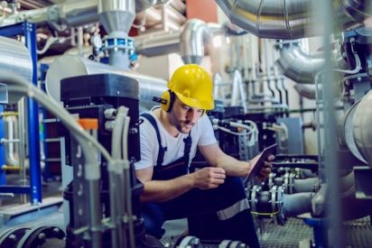 Factory worker wearing safety gear reviews live production metrics on a tablet beside industrial equipment as part of short-interval control process.