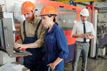 Factory operators and supervisors gathered during a Gemba walk, reviewing visual work instructions and one point lessons to support lean manufacturing, continuous improvement, safety training, and process standardization.