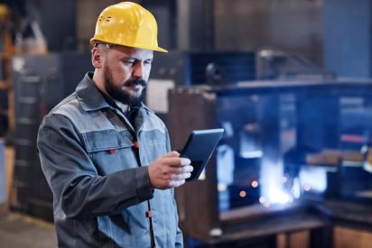 A manufacturing operator uses a tablet beside a production line, demonstrating paperless manufacturing, digital work instructions, and real-time shop floor operations.