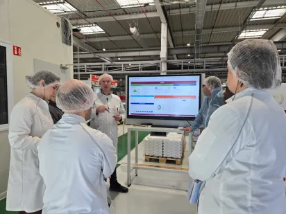 A team of manufacturing workers gathered around a digital display board during a daily huddle meeting to review operational KPIs, an example of a lean daily management system in practice.